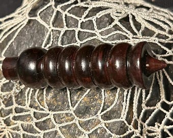 Op de afbeelding: Een donkerbruine houten flesstopper met een reeks afgeronde, geribbelde secties en een puntig uiteinde. De stopper staat tegen een getextureerde, donkere achtergrond met een witte, netachtige overlay.