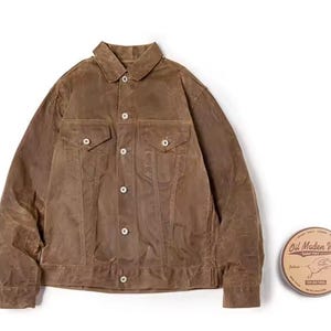 May include: A brown, button-up jacket with two chest pockets and two side pockets. The jacket has a classic collar and is made of a waxed material. A round tin of "Oil Maden Wax" is next to the jacket.