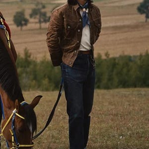 May include: A person wearing a brown jacket, white shirt, and dark blue jeans stands in a field next to a horse. The person is also wearing a blue patterned scarf. The text on the image reads: 174cm/70kg/L.