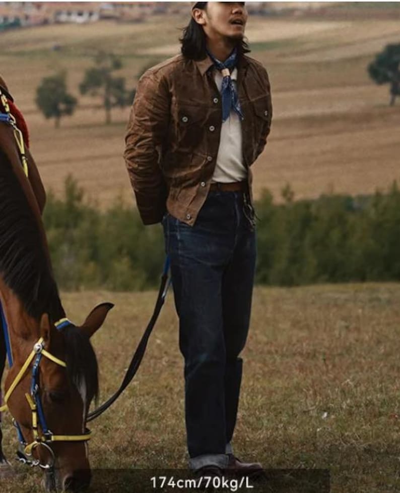 May include: A person wearing a brown jacket, white shirt, and dark blue jeans stands in a field next to a horse. The person is also wearing a blue patterned scarf. The text on the image reads: 174cm/70kg/L.