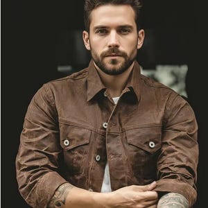 May include: A brown jacket with two chest pockets and button fastenings. The jacket has a collared neckline and is worn over a white shirt. The sleeves are long and buttoned at the cuffs. The jacket appears to be made of a durable material.