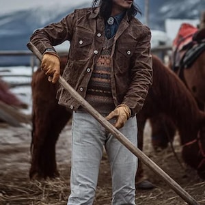 May include: A brown jacket with button closures, worn over a patterned sweater and denim shirt. The person is wearing a hat, sunglasses, and gloves, holding a wooden pole. The image includes the text "170cm/67kg/M".