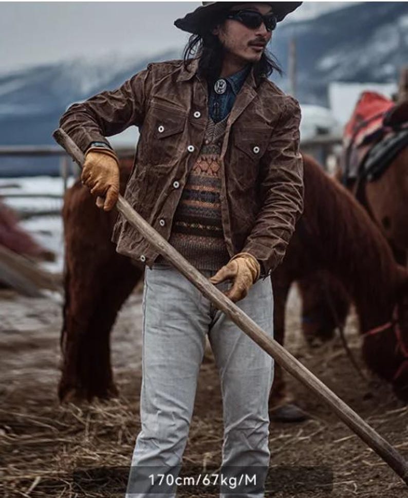 May include: A brown jacket with button closures, worn over a patterned sweater and denim shirt. The person is wearing a hat, sunglasses, and gloves, holding a wooden pole. The image includes the text "170cm/67kg/M".