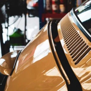 May include: Close-up of a vintage tan car's body, showcasing the hood, side mirror, and air vent. The car's glossy surface reflects the surrounding environment. The image highlights the car's classic design and color.