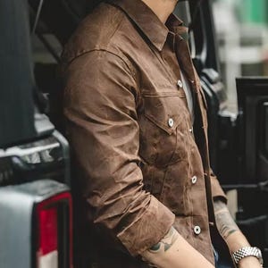 May include: A brown jacket with a collared neckline and two chest pockets. The jacket has white buttons and long sleeves. The fabric has a textured appearance. The jacket is worn over a white shirt.