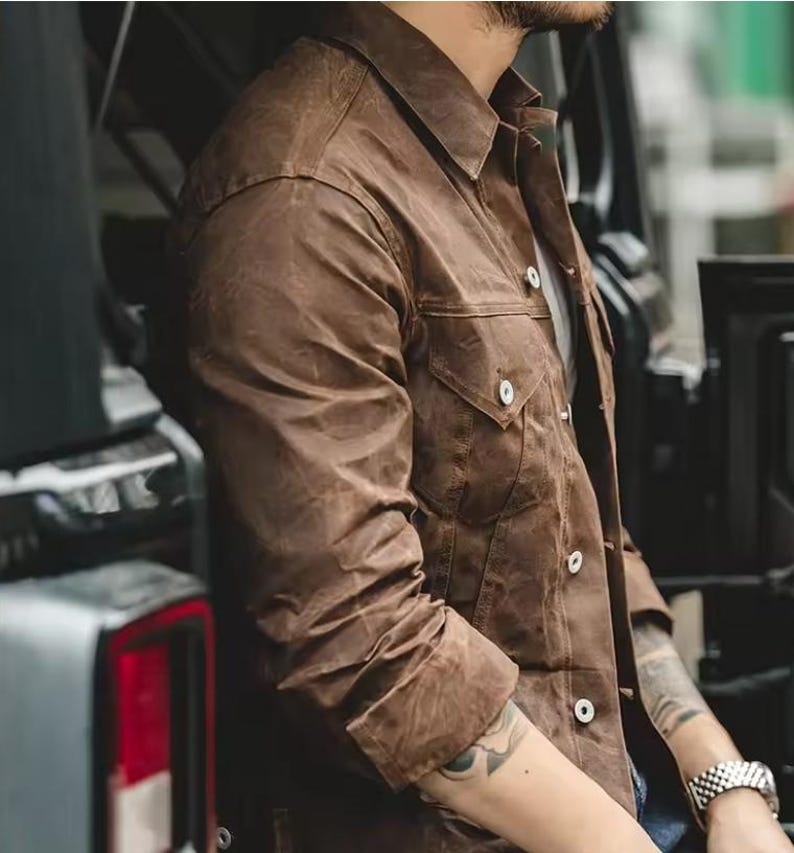 May include: A brown jacket with a collared neckline and two chest pockets. The jacket has white buttons and long sleeves. The fabric has a textured appearance. The jacket is worn over a white shirt.