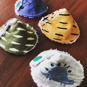 Puede incluir: Cuatro baberos de tela con diferentes patrones. Los baberos son en forma de cono y tienen un borde blanco y peludo. Los patrones incluyen un diseño de nubes azul y blanco, un diseño de rayas amarillas y negras, un diseño de rayas verdes y blancas y una tela blanca con un diseño de dinosaurio azul.