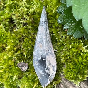 May include: A teardrop-shaped stone pendant featuring a polished surface with a marbled pattern in shades of gray, black, and white. The stone has a smooth, elongated form that tapers to a point at the top and is rounded at the base.