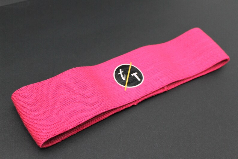Puede incluir: Banda de resistencia de tela rosa con un logotipo circular negro que dice "t/t" en blanco.