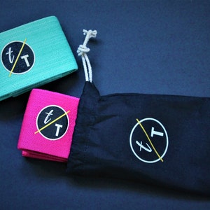 Puede incluir: Dos bandas de resistencia de tela en color turquesa y rosa con un logotipo negro y amarillo que dice "t/T". Las bandas est&aacute;n enrolladas y se encuentran sobre una bolsa de tela negra con cord&oacute;n y el mismo logotipo.