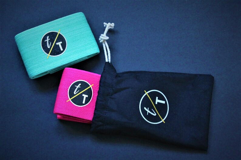 Puede incluir: Dos bandas de resistencia de tela en color turquesa y rosa con un logotipo negro y amarillo que dice "t/T". Las bandas est&aacute;n enrolladas y se encuentran sobre una bolsa de tela negra con cord&oacute;n y el mismo logotipo.