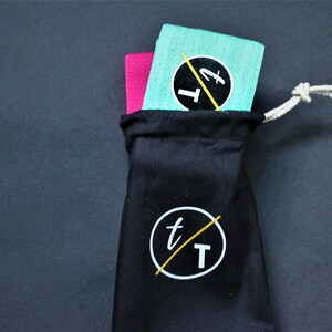 Puede incluir: Dos bandas de resistencia, una rosa y una turquesa, est&aacute;n dobladas y colocadas dentro de una bolsa de cord&oacute;n negro con un logotipo blanco que dice "t/T".