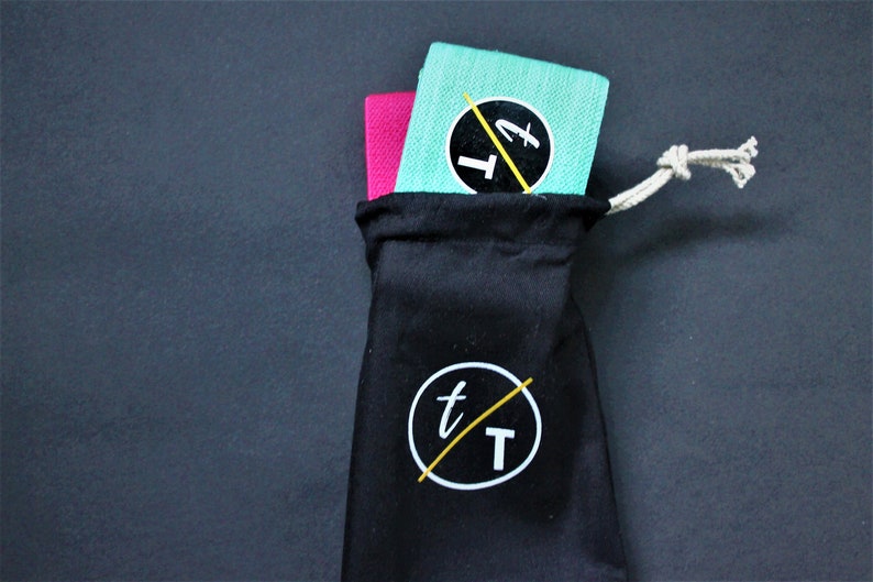 Puede incluir: Dos bandas de resistencia, una rosa y una turquesa, est&aacute;n dobladas y colocadas dentro de una bolsa de cord&oacute;n negro con un logotipo blanco que dice "t/T".