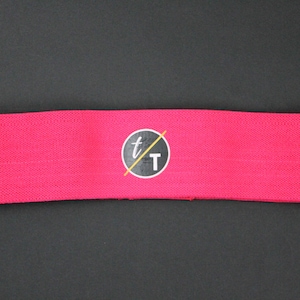 Puede incluir: Banda de resistencia de tela rosa con un logotipo circular negro que dice "t/T".