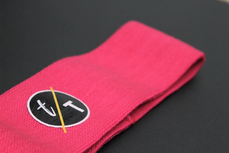 Puede incluir: Banda de resistencia de tela rosa con un logotipo circular negro que dice "T/T" en letras blancas con una l&iacute;nea amarilla que lo atraviesa.