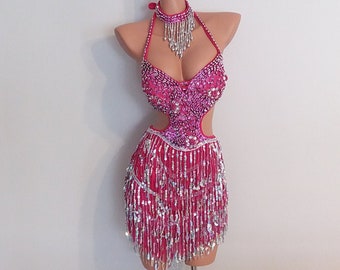 May include: A pink and silver sequined dance costume with fringe. The costume has a halter top with a plunging neckline and a skirt with a high slit.