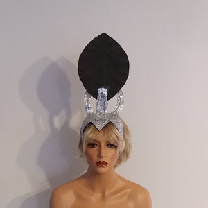 May include: A silver and black headpiece with a large, round, black top and a silver, pointed crown. The headpiece is on a white mannequin head.