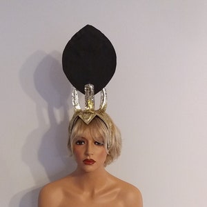 May include: A gold and black headpiece with a large black feather on top. The headpiece is worn by a mannequin with blonde hair.