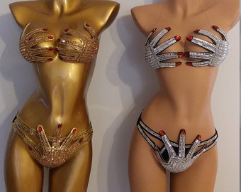 May include: Two mannequins display bejeweled lingerie sets. One is gold with red accents, the other silver with red accents. The bra and panty sets are designed to resemble hands. The mannequins are on gold stands.