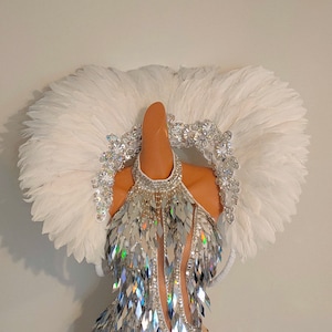 Puede incluir: Un body plateado con lentejuelas iridiscentes en forma de diamante y un tocado de plumas blancas. El disfraz presenta un cuello halter y cortes. El tocado es una gran disposición circular de plumas blancas. Pedrería y adornos plateados adornan el escote y el tocado.