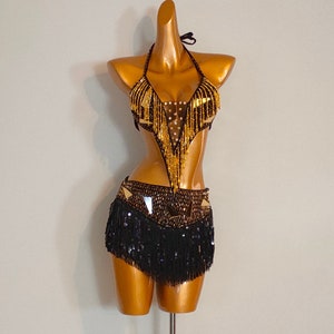 Puede incluir: Un conjunto de disfraz negro y dorado con cuentas, con un top halter y una falda. El top tiene un diseño de flecos y la falda tiene un fleco de lentejuelas.