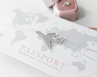 May include: A silver airplane charm with the date "A.M. 23.06.2022" engraved on it hangs from a silver chain on a white card with a world map and the text "PASSPORT WEDDING INVITATION".