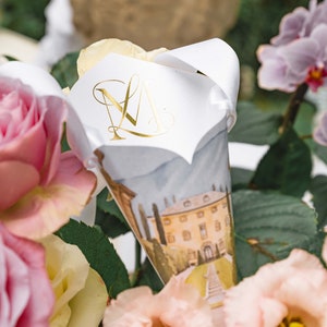 May include: A white paper cone with a gold monogram "M" on the front. The cone is decorated with a watercolour illustration of a building with a garden and a walkway. The cone is surrounded by pink and yellow flowers.