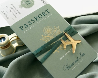 May include: A green passport-style wedding invitation with gold lettering that reads "Passport Wedding Invitation". A gold airplane charm is attached to a green ribbon tied around the invitation. The text "20th April 20 Victoria and Sean Como Lake, Italy" is printed on the invitation.