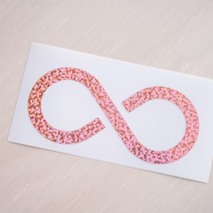 May include: A white rectangular sticker featuring a pink and gold glitter infinity symbol. The symbol is centered on the sticker, with a light-colored background.