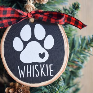 May include: A black and white wooden Christmas ornament with a white paw print and a small red heart. The ornament is decorated with a red and black buffalo plaid ribbon and the name "WHISKIE" is written in white on the bottom.