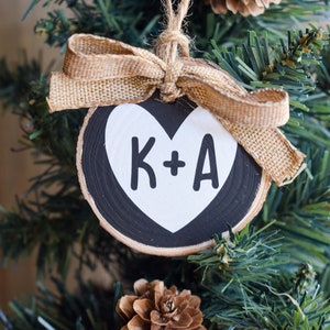 May include: A black and white wooden ornament with a heart shape and the text "K+A" on it. The ornament is tied with a brown burlap ribbon.