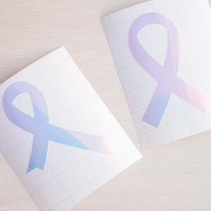 May include: Two sheets of clear transfer paper with iridescent blue ribbon designs. The ribbons are in the shape of awareness ribbons.