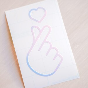 May include: A white sticker featuring a hand gesture forming a heart shape, with a heart symbol above. The design has a gradient effect, transitioning from pink to blue. The sticker is on a white background.