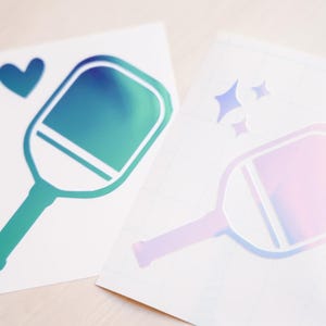 May include: Two iridescent vinyl stickers of a pickleball paddle. One paddle is teal and green, the other is pink and purple. The teal and green paddle has a heart above it. The pink and purple paddle has three stars above it.