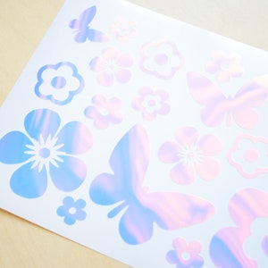 May include: A sheet of iridescent stickers featuring butterflies and flowers in a pastel color scheme. The stickers are perfect for adding a touch of whimsy to journals, laptops, or other surfaces.