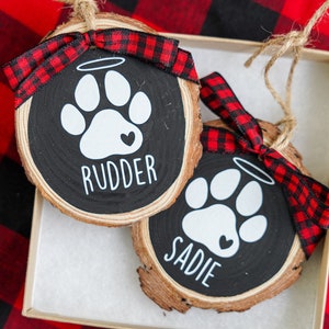 May include: Two wooden ornaments with black painted surfaces and white paw prints. Each ornament has a red and black buffalo plaid ribbon and a white halo above the paw print. The ornaments are personalized with the names "Rudder" and "Sadie".