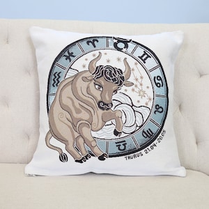 May include: White throw pillow featuring a brown bull illustration within a blue circle containing zodiac symbols and the text "Taurus 21.04-20.05".