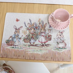 Op de afbeelding: Een decoratieve placemat met een illustratie van konijnen in een tuin, elk met manden met bloemen. Een roze kop en schotel staan op de placemat, die op een lichte houten tafel ligt.