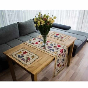 May include: A wooden table with two beige table runners featuring a colourful butterfly and floral design. The table runners are placed on a wooden table with a vase of yellow flowers in the centre.