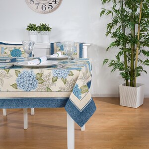 Puede incluir: Una mesa blanca con un mantel floral azul y blanco. La mesa está puesta con dos cubiertos, cada uno con un plato blanco, una servilleta blanca y un vaso transparente. Hay un reloj en la pared detrás de la mesa.
