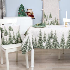 Pode incluir: Mesa de jantar branca com uma toalha de mesa branca com um padrão de pinheiros verdes. Duas cadeiras brancas com almofadas com padrão de pinheiros verdes são colocadas na mesa. Uma pequena árvore de Natal com enfeites está no fundo.