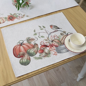 Può includere: Un tovaglietta beige con un ricamo di zucche, zucche, funghi e un pettirosso appollaiato su un ramo. La tovaglietta è su un tavolo di legno con una tazza e un piattino bianchi.