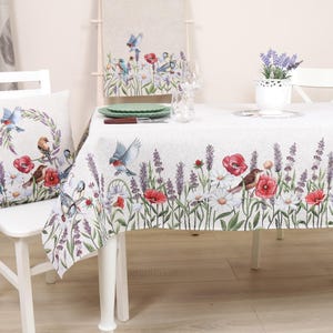 May include: A white tablecloth, pillow, and runner with a floral and bird design. The design features red poppies, white daisies, purple lavender, and blue birds. A white chair and table are in the image.