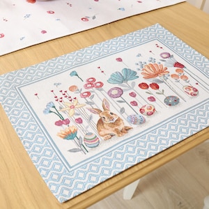 Può includere: Un sottopiatto rettangolare con un bordo geometrico azzurro e un disegno centrale con un coniglio marrone, fiori colorati e uova di Pasqua. Il sottopiatto è posto su un tavolo di legno, con un runner abbinato visibile sullo sfondo.