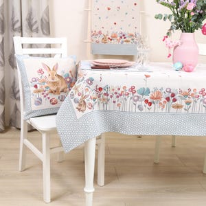 Könnte beinhalten: Weißer Tisch mit einer floralen Tischdecke mit einem Rand aus Hasen und Eiern. Die Tischdecke hat einen blau-weißen geometrischen Muster am unteren Rand. Ein weißer Stuhl mit einem Kissen mit einem Hasen und Eiern steht am Tisch.