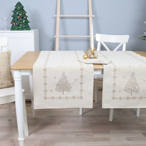 Peut inclure: Deux chemins de table beiges avec des sapins de Noël brodés en or et des flocons de neige sur une table en bois blanc. Les chemins de table sont dressés pour un dîner de fête avec une chaise blanche et un coussin avec un sapin de Noël brodé en or.