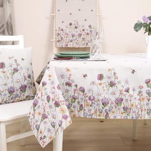 May include: White tablecloth with a floral pattern featuring daisies, bees, and other wildflowers. The tablecloth is set on a white table with two white chairs and a vase of flowers.