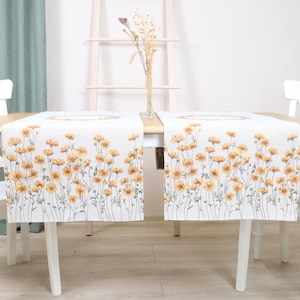 Puede incluir: Dos caminos de mesa blancos con un diseño floral de flores amarillas y tallos verdes. Los caminos de mesa están colocados sobre una mesa de madera clara, con sillas blancas a ambos lados. Un jarrón con flores secas está en el centro.