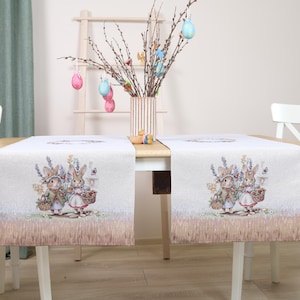 Puede incluir: Dos caminos de mesa con temática de Pascua con una ilustración de conejos. Los caminos de mesa beige tienen una impresión detallada de conejos con cestas de flores. La mesa está decorada con huevos de colores y un jarrón con ramas.