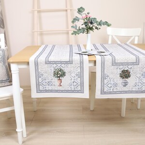 Pode incluir: Dois caminhos de mesa brancos com um padrão floral cinza e branco. Os caminhos de mesa estão sobre uma mesa branca com uma cadeira branca e uma escada branca no fundo.